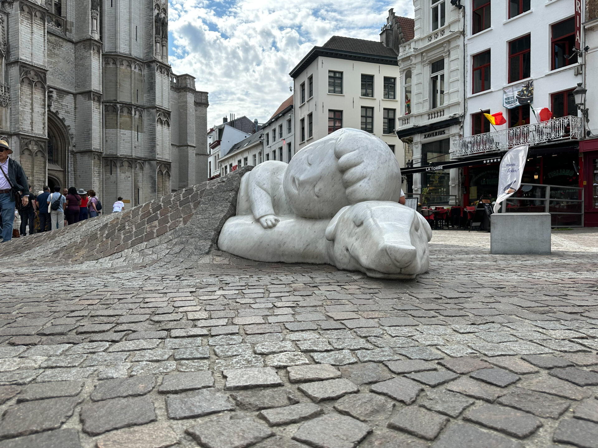 Direkt an der Liebfrauenkathedrale findest du die Figuren Nello und Patrasche, die in liebevoller Umarmung auf den Pflastersteinen liegen. Die Skulptur des Künstlers Batist Vermeulen zeigt die Protagonisten des englischen Romans "A Dog of Flanders", der 1872 veröffentlicht wurde und vor allem außerhalb Belgiens Berühmtheit erlangte. Im Roman schließt der arme Waisenjunge Nello Freundschaft mit dem Straßenhund Patrasche. Zusammen erkunden sie Antwerpen, besuchen die Liebfrauenkathdrale und bewundern die Gemälde von Rubens.